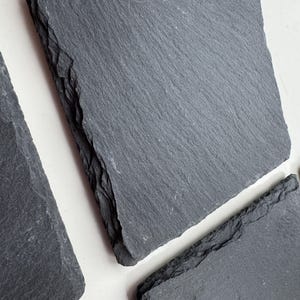 May include: A set of square slate coasters featuring a dark grey colour and a natural, rough-hewn texture on the edges. The surface of each coaster is flat with a subtle, layered grain pattern, showcasing the authentic, unpolished look of stone.