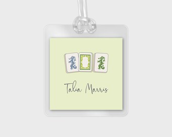 May include: A light yellow luggage tag with a white border. The tag features three green and blue mahjong tiles with dragons. The text "Talia Marris" is printed in black below the tiles.