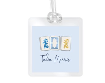May include: A light blue luggage tag with three white mahjong tiles featuring blue and gold dragons. The tag has the name "Talia Marris" written in black cursive.