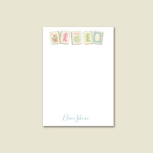 May include: A white notepad with a blue and green border of mahjong tiles. The tiles have Chinese characters and the word "Joker" on them. The notepad has the name "Elaine Johnson" written in blue cursive at the bottom.