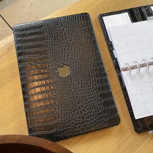 May include: A black laptop cover with a textured crocodile skin pattern and a central cutout for an Apple logo, positioned next to an open black planner with a white monthly calendar grid, silver disc binding, and a black pen resting on the cover.