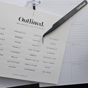 May include: A planner with a black cover and white stickers. The stickers are labelled with categories such as "Projects", "Home", and "Expenses". A black tweezer with the word "Outlined" in gold is holding a sticker. The planner is open to a 2025 calendar.