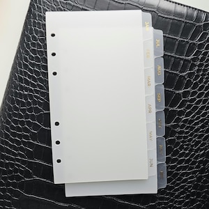 May include: White planner inserts with monthly tabs labeled January through December in gold lettering. The inserts have pre-punched holes for binding. The background is a black, textured surface.