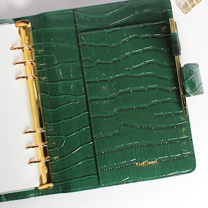 May include: Green faux-leather planner with a crocodile skin texture. The planner has gold-colored metal rings and a pen holder. The word "Outlined" is printed in gold on the front. A small notepad and gold binder clips are also visible.