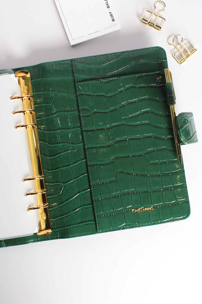 May include: Green faux-leather planner with a crocodile skin texture. The planner has gold-colored metal rings and a pen holder. The word "Outlined" is printed in gold on the front. A small notepad and gold binder clips are also visible.