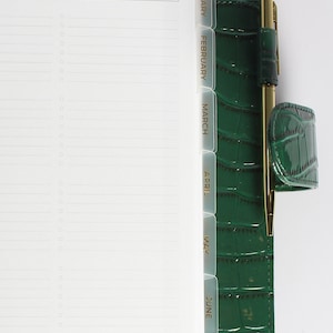 May include: A green faux leather planner with gold accents and clear dividers labeled January through June. The planner has a pen holder and a section labeled "Financial Goals" and "Action Plan".