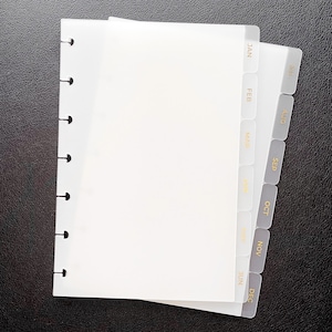 Op de afbeelding: Een witte planner met maandelijkse tabbladen met de maanden januari tot en met december. De planner heeft een spiraalbinding aan de linkerkant. De tabbladen zijn grijs met gouden letters. De planner is open en toont lege pagina's.