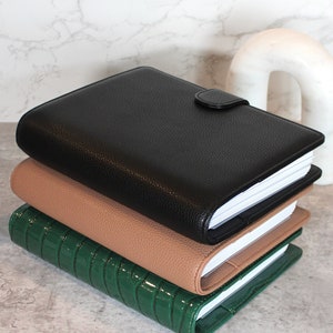 May include: Three stacked leather-bound journals in black, tan, and green. The black journal has a textured finish and a snap closure. The tan journal is smooth, and the green journal has a glossy, crocodile-like texture. The pages are white.