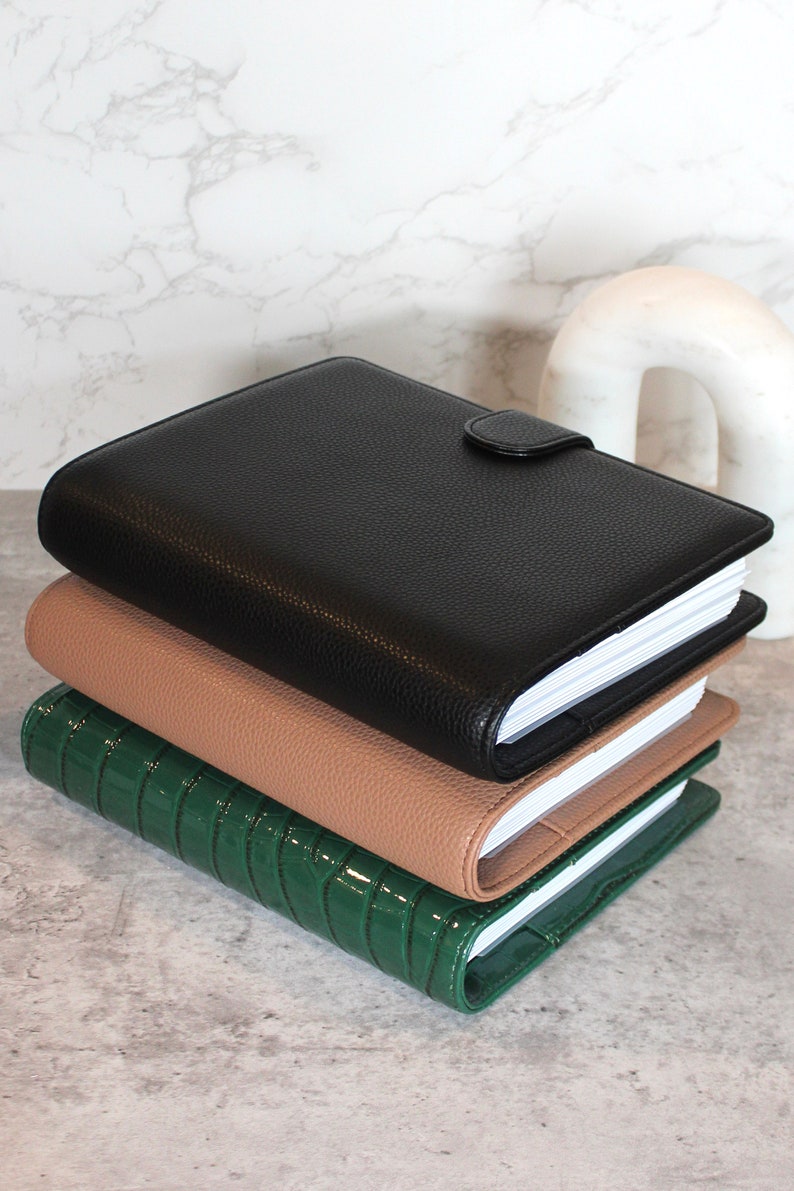 May include: Three stacked leather-bound journals in black, tan, and green. The black journal has a textured finish and a snap closure. The tan journal is smooth, and the green journal has a glossy, crocodile-like texture. The pages are white.
