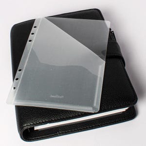 Puede incluir: Un planificador de cuero negro con una página de bolsillo de plástico transparente en la parte superior. La página de bolsillo tiene un diseño diagonal y cubre parcialmente el planificador. El planificador está cerrado y tiene un cierre de correa.