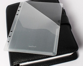 May include: A black leather planner with a clear plastic pocket page on top. The pocket page has a diagonal design and is partially covering the planner. The planner is closed and has a strap closure.