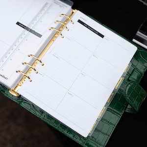May include: A green leather planner with gold accents, open to a weekly spread. The planner features gold rings, a ruler, and month tabs. The pages are white with black text, and the cover has a crocodile texture.
