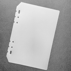 May include: A white plastic sheet with a rounded corner and holes along the left edge. The sheet is lying on a black textured surface.