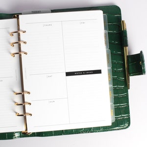 May include: Green faux-leather planner with gold-colored accents. The planner is open to a weekly spread with lined pages and day labels. A gold pen is secured in a loop. Tabs mark months. The word "TODAY" is visible.