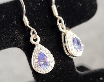 Può includere: Orecchini in argento con una pietra preziosa a forma di goccia di colore viola chiaro, circondata da piccole pietre trasparenti. Gli orecchini sono esposti su un espositore di gioielli in velluto nero.