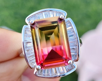 May include: A silver ring with a large, rectangular, multi-colored gemstone. The gemstone is a gradient of yellow, orange, and red. The ring is set with smaller, clear gemstones around the main stone.