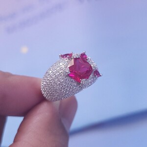 Può includere: Un anello d'argento con una grande pietra rossa circondata da pietre rosse più piccole e una fascia di piccole pietre bianche.