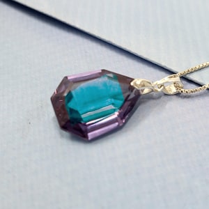 May include: A silver necklace featuring a pendant with a faceted, gemstone-like design. The pendant has a teal center and is framed by a purple hue. The chain is a delicate silver, and the pendant is the focal point.