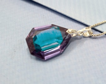 May include: A silver necklace featuring a pendant with a faceted, gemstone-like design. The pendant has a teal center and is framed by a purple hue. The chain is a delicate silver, and the pendant is the focal point.