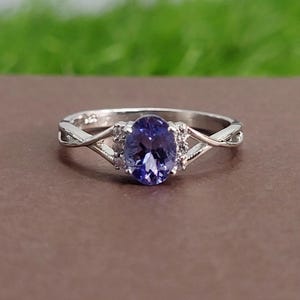 May include: A silver ring with a large, oval, deep blue gemstone. The gemstone is set between smaller, clear stones, and the band has a delicate, twisted design. The ring is shown against a brown surface with a blurred green background.
