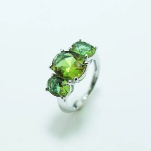 Puede incluir: Un anillo de plata con tres piedras verdes. La piedra central es de un verde claro brillante, mientras que las otras dos son de un tono verde más oscuro. La banda del anillo es de plata pulida y las piedras están engarzadas con pequeñas garras.