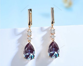 May include: A pair of rose gold earrings with a teardrop-shaped, color-changing gemstone. The earrings feature a small cluster of clear crystals and a lever back closure. The gemstones transition from purple to teal.