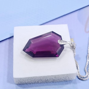 Könnte beinhalten: Ein facettierter, tiefvioletter Edelsteinanhänger mit silberner Öse und Kette. Der Anhänger hat eine geometrische Form mit mehreren Facetten, die das Licht reflektieren. Die Kette ist eine filigrane silberne Boxkette. Der Anhänger liegt auf einem weißen Schaumstoffpad.
