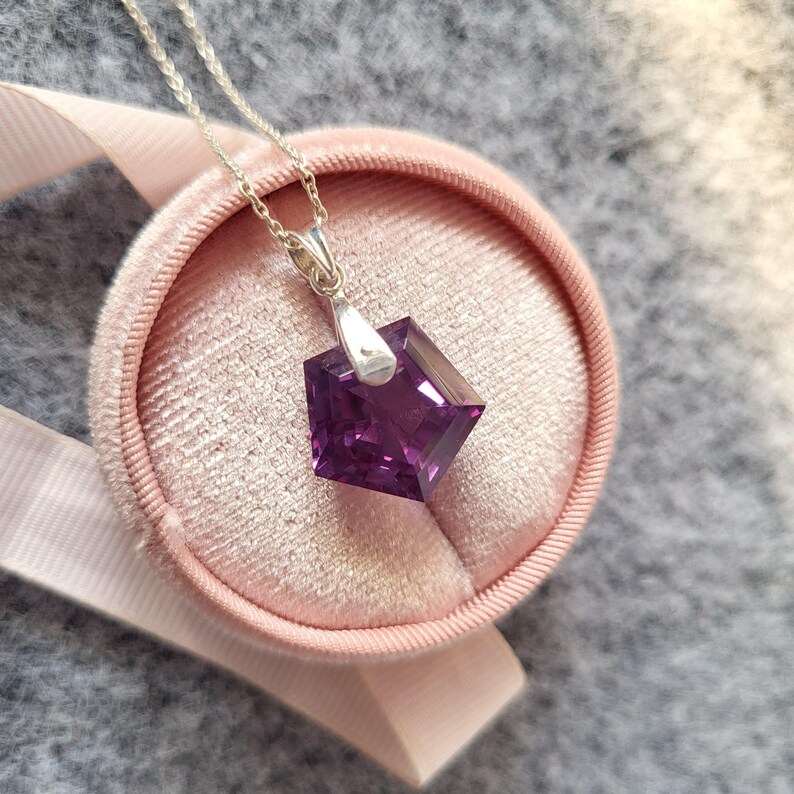 May include: A silver necklace with a square, faceted, deep purple pendant. The pendant is displayed on a pink velvet jewelry box with a matching ribbon. The necklace chain is delicate and the pendant is the focal point.