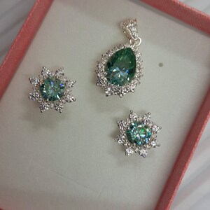 May include: A set of jewelry featuring a teardrop-shaped pendant and matching earrings. The pendant and earrings have a green gemstone surrounded by a halo of smaller white gemstones.