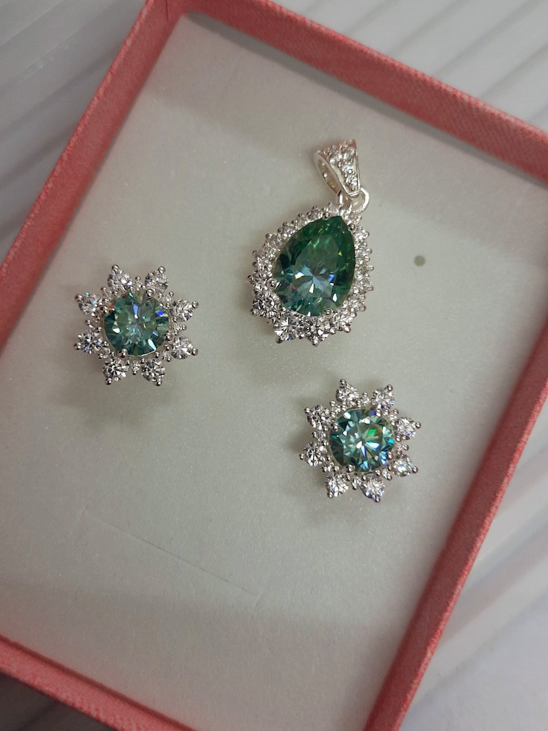 May include: A set of jewelry featuring a teardrop-shaped pendant and matching earrings. The pendant and earrings have a green gemstone surrounded by a halo of smaller white gemstones.
