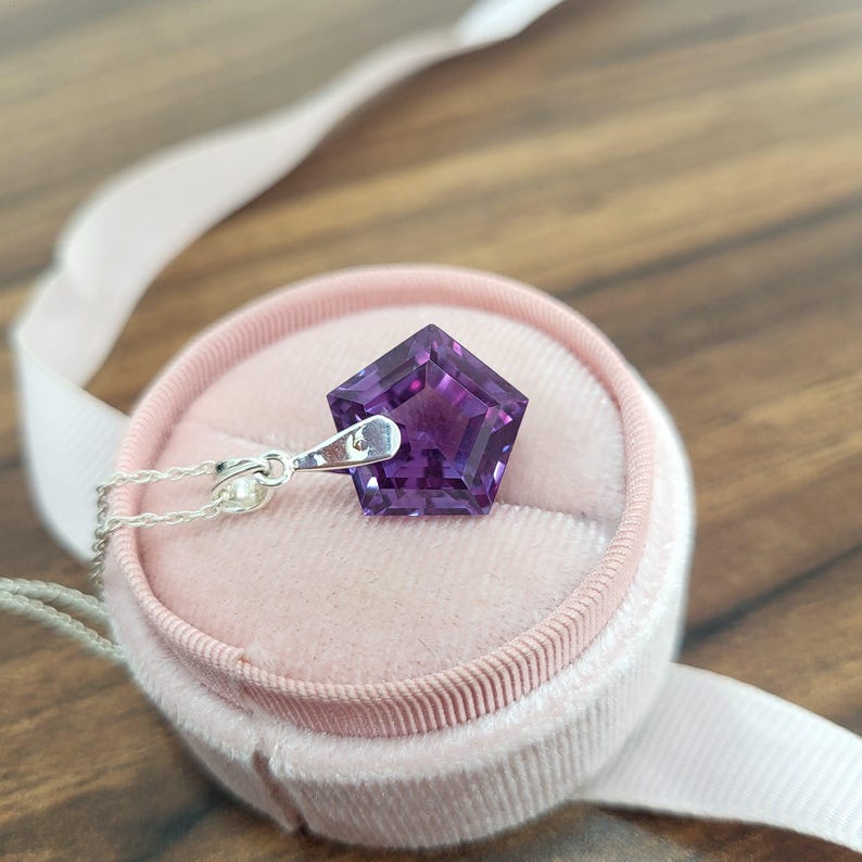 May include: A pentagon-shaped, deep purple gemstone pendant on a silver chain. The pendant is set in a light pink velvet jewelry box with a white ribbon. The gemstone has a faceted surface, catching the light.