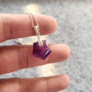 May include: A silver necklace with a geometric, faceted, deep purple pendant. The pendant is a five-sided shape, catching the light. The chain is delicate and the pendant is held by a silver bail.