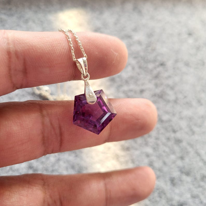 May include: A silver necklace with a geometric, faceted, deep purple pendant. The pendant is a five-sided shape, catching the light. The chain is delicate and the pendant is held by a silver bail.