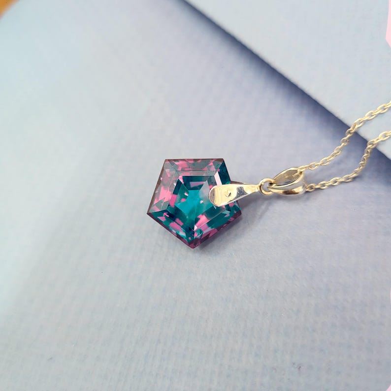 May include: A silver necklace featuring a pendant with a geometric, pentagon-shaped gemstone. The gemstone displays a blend of teal and magenta hues. The chain is delicate and the pendant is attached with a small clasp.