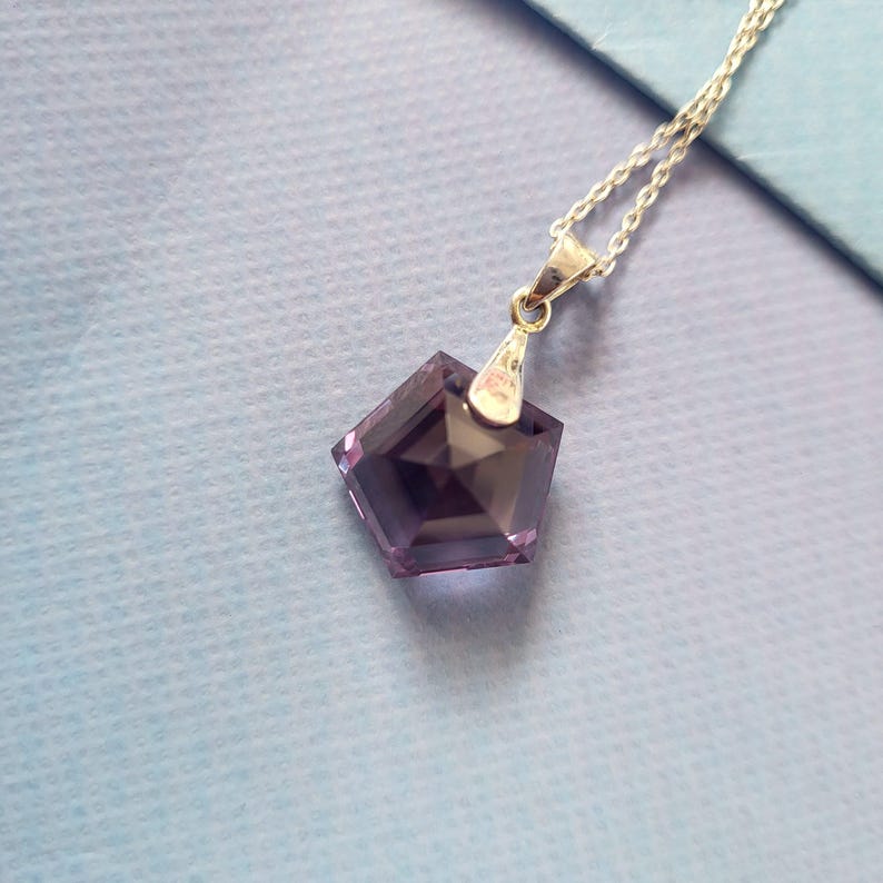May include: A silver chain necklace with a faceted, pentagon-shaped, purple pendant. The pendant has a silver bail and is suspended from a delicate silver chain. The necklace is displayed on a light blue surface.