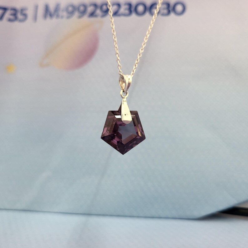 May include: A silver necklace with a pentagon-shaped, dark purple pendant. The pendant is faceted, catching the light. The chain is delicate, and the pendant hangs from a small silver bail. The background is a light blue.