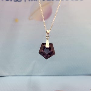 May include: A silver necklace featuring a dark purple, pentagon-shaped pendant. The pendant is faceted, catching the light and displaying a deep, rich color. The chain is delicate, and the pendant hangs from a small silver bail.