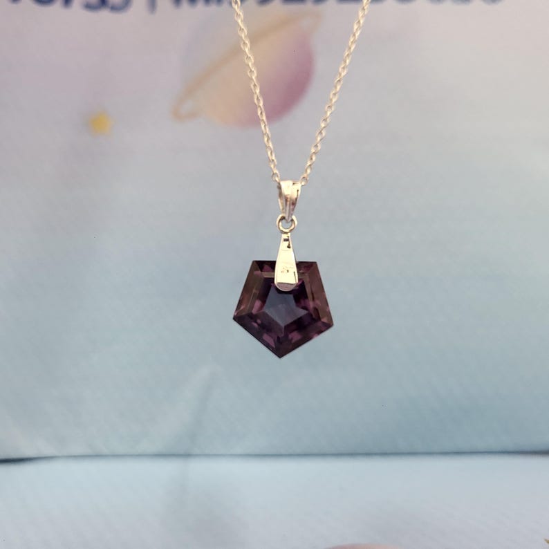 May include: A silver necklace featuring a dark purple, pentagon-shaped pendant. The pendant is faceted, catching the light and displaying a deep, rich color. The chain is delicate, and the pendant hangs from a small silver bail.