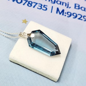 May include: A blue gemstone pendant with a silver chain. The pendant is a geometric shape with a pointed end. The gemstone is a light blue color and is set in a silver setting. The pendant is displayed on a white surface.