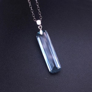 Puede incluir: Un collar con un colgante rectangular azul claro y una cadena plateada. El colgante es un cristal claro y alargado con un diseño facetado. El collar se muestra sobre un fondo negro.