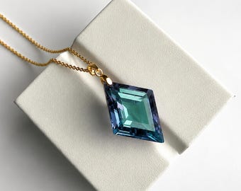 May include: A gold chain necklace featuring a large, faceted, diamond-shaped pendant. The pendant displays a color-shifting gradient transitioning from deep blue to vibrant teal, with sharp geometric edges and a polished, reflective surface finish.