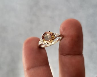 May include: A silver ring featuring a hexagonal, amber-colored gemstone as the centerpiece. The ring is accented with smaller, clear gemstones on either side of the main stone, creating a delicate and elegant design. The ring is held up by a finger.