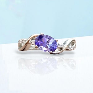 Può includere: Un anello d'argento con una grande pietra preziosa viola a taglio ovale. La fascia presenta un design intrecciato con piccole pietre chiare. L'anello è posto su una superficie bianca con sfondo azzurro.