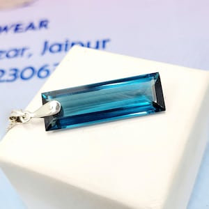 May include: A rectangular, teal gemstone pendant with a silver bail and chain. The faceted gemstone displays clarity and colour. The pendant is presented on a white surface.