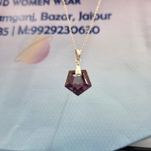 May include: A silver necklace with a geometric, dark purple pendant. The pendant is a faceted, pentagon shape with a silver bail. The chain is delicate and hangs from the bail. The background is a light blue and white.