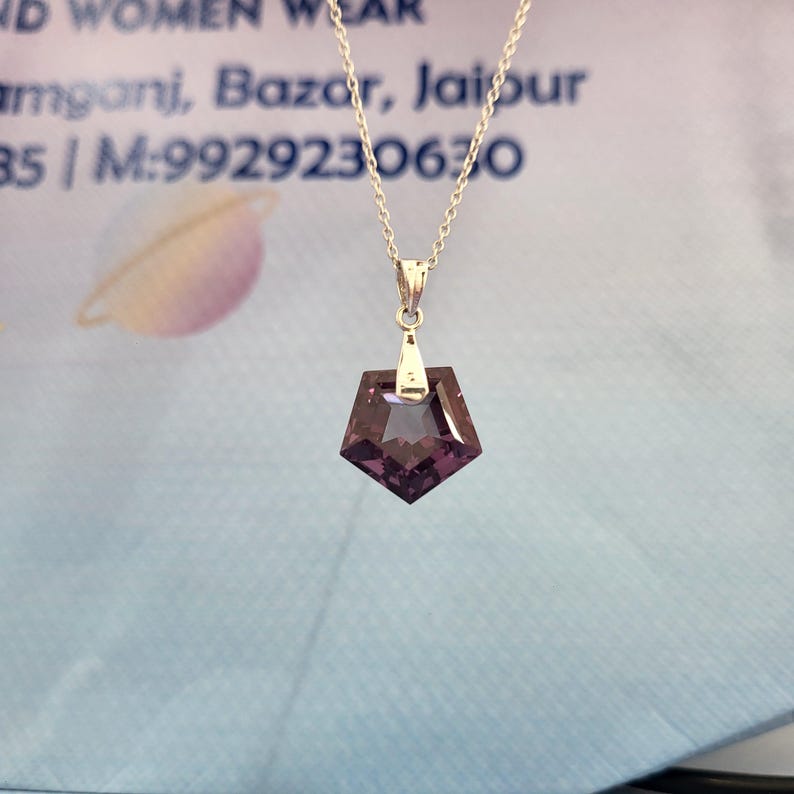 May include: A silver necklace with a geometric, dark purple pendant. The pendant is a faceted, pentagon shape with a silver bail. The chain is delicate and hangs from the bail. The background is a light blue and white.