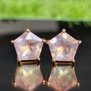 Op de afbeelding: Een paar oorknopjes met vijfhoekige, lichtroze gefacetteerde edelstenen gezet in roségouden metalen pootjes. De oorbellen staan naast elkaar en vertonen een duidelijke reflectie van hun geometrische vorm op het oppervlak eronder.