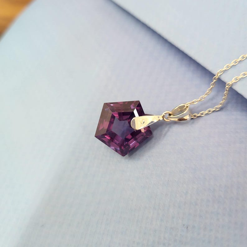 May include: A faceted, pentagon-shaped pendant necklace with a deep purple gemstone. The pendant is suspended from a delicate silver chain. The gemstone has a polished, reflective surface. The necklace is displayed on a light blue surface.