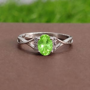 Puede incluir: Un anillo de plata con una piedra preciosa ovalada de color verde lima en el centro, flanqueada por pequeñas piedras claras a cada lado. La banda tiene un diseño dividido y retorcido, y está marcada con 925, indicando plata de ley.