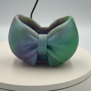 May include: A small, hollow container shaped like a bow, featuring a gradient of blended purple, blue, and green colors. The surface has a subtle, fine-lined texture throughout, with a central vertical band detail connecting the two sides.